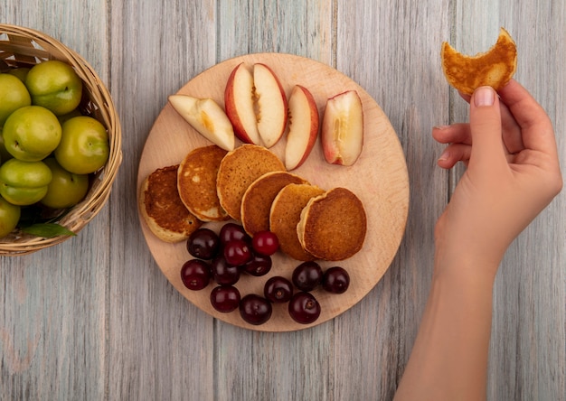 Top view of female hand holding bitten pancake and pancakes with cherries and sliced peach on cutting board with basket of plums on wooden background