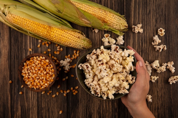 Top view of female hand holding a bowl of popcorns with corn kernels on a wooden bowl with fresh corns on a wooden table