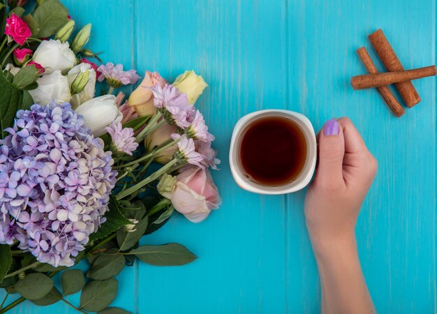 Top view of female hand holding a cup of tea with cinnamon stick fresh flowers isolated on a blue wooden background