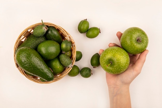 Top view of female hand holding fresh apples with feijoas and avocados on a bucket on a white surface