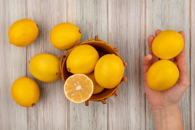 Top view of female hand holding lemons with lemons on a bucket on a grey wooden surface