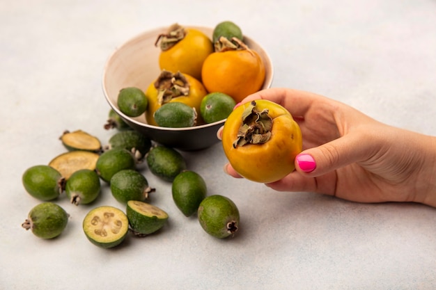 Top view of female hand holding an orange ripe persimmon fruit with feijoas and persimmons isolated on a grey surface