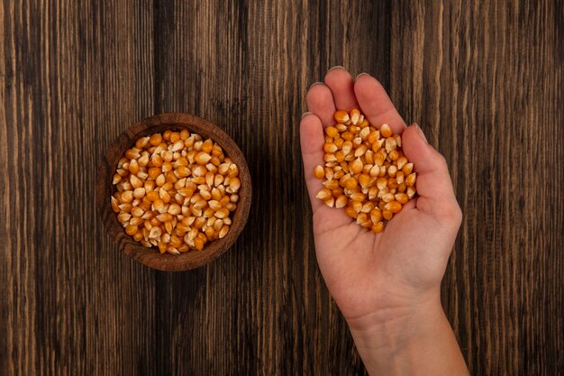 Top view of female hand holding organic corn kernels with corn kernels on a wooden bowl on a wooden table