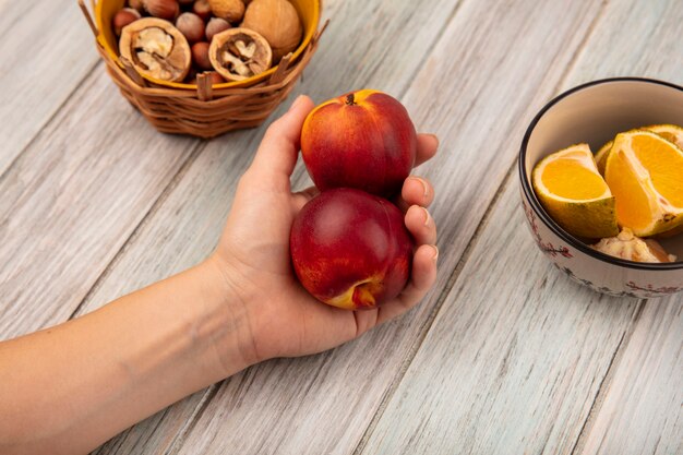 Вид сверху женской руки, держащей персики с орехами на ведре с мандаринами на миске на серой деревянной поверхности