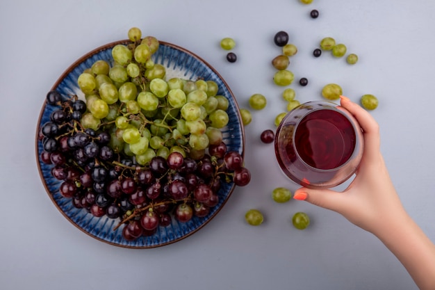 Vista superiore della mano femminile che tiene il vino rosso nel bicchiere di vino e uva nel piatto e su sfondo grigio