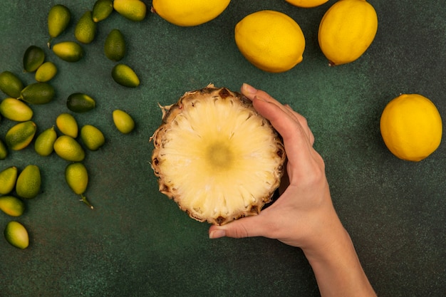 Вид сверху женской руки, держащей сладкую половину ананаса с лимонами и кинканами, изолированными на зеленой стене