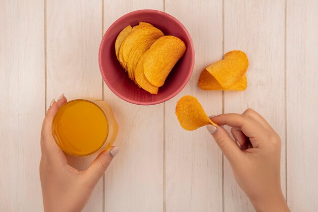 Top view of female hand holding tasty crispy chips with a glass of orange juice on a beige wooden table
