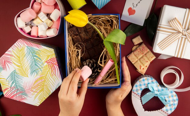 Top view of female hand putting marshmallow into a present box with yellow color tulip flower , dark chocolate bar , cone and straw on dark red table with a box filled with marshmallow