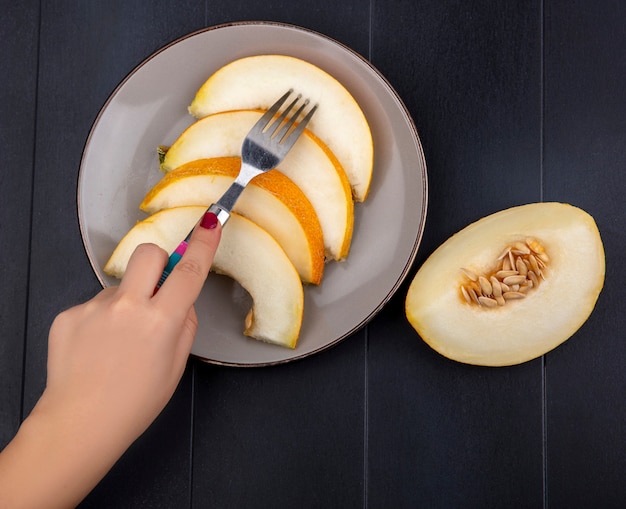 Top view of female hand taking slice of melon with fork on plate on black