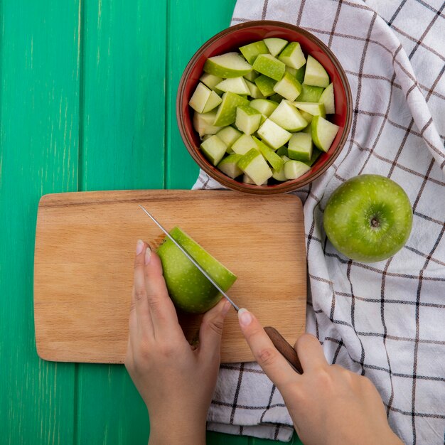 Вид сверху на женские руки, рубящие зеленые яблоки на деревянной кухонной доске на красной миске нарезанных яблок и ткани