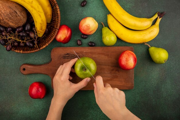 Top view of female hands cutting apple with knife on cutting board and banana pear cococnut grape peach on green background