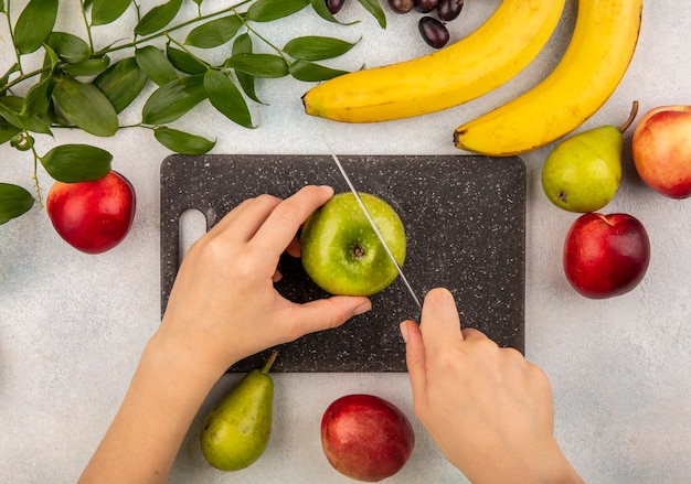 Vista superiore delle mani femminili che tagliano la mela con il coltello sul tagliere e la pesca della banana della pera dell'uva con i fogli su fondo bianco