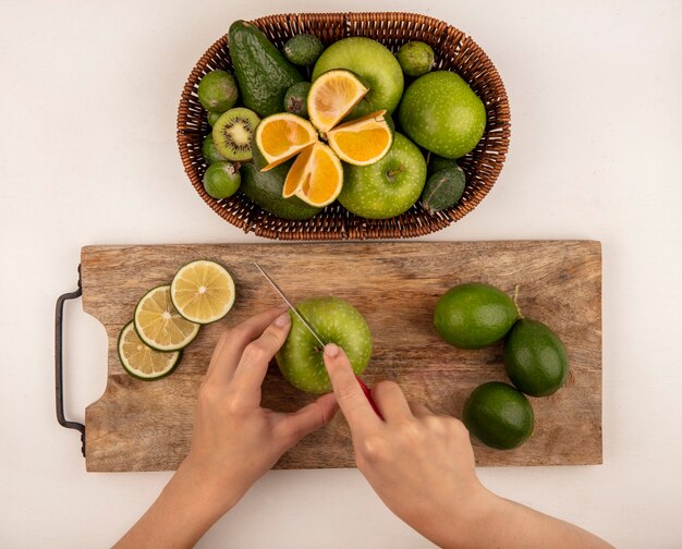 Top view of female hands cutting an apple on a wooden kitchen board with knife with a bucket of green apples kiwi feijoas and limes on a white wall