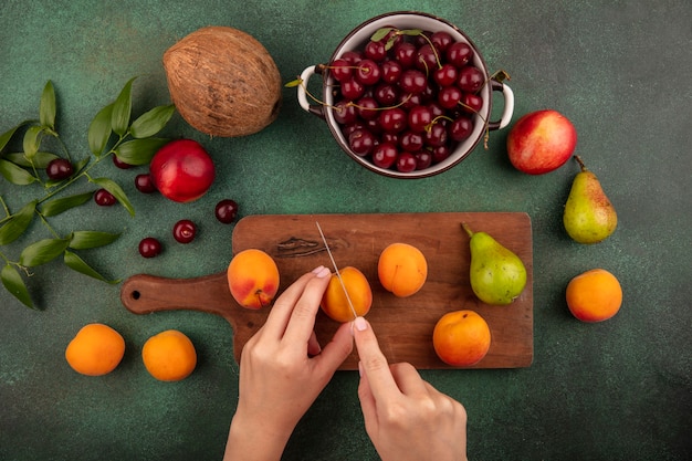 Top view of female hands cutting apricot with knife and pear on cutting board and cherries in bowl with peaches coconut and leaves on green background