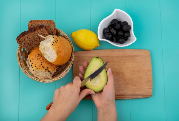 Top view of female hands cutting avocado with knife on wooden kitchen board with a bucket of breads with black olives on white bowl on blue