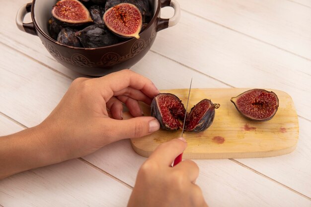 Top view of female hands cutting black figs on a wooden kitchen board with knife on a white wooden wall