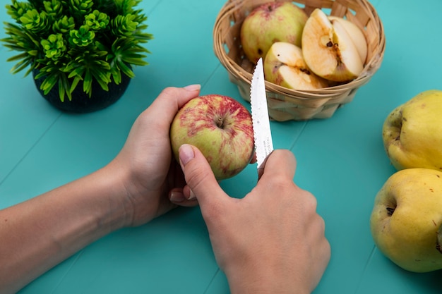 Вид сверху женских рук, режущих свежее яблоко ножом на синем фоне