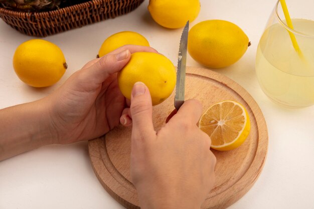 Top view of female hands cutting fresh lemon on a wooden kitchen board with knife with lemons and lemon juice on a white surface