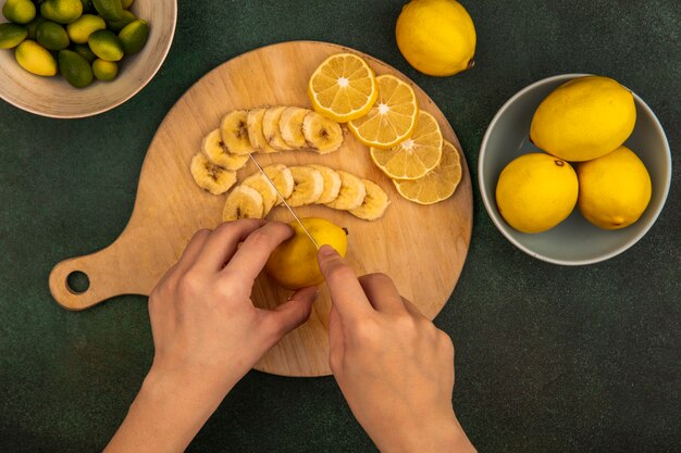 Top view of female hands cutting fresh lemons on a wooden kitchen board with knife with kinkans on a bowl on a green surface