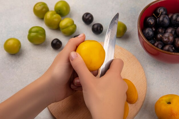 Top view of female hands cutting fresh peach with knife on a wooden kitchen board with knife with sloes on a red bowl on a white background