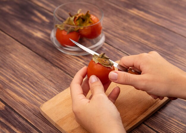 Top view of female hands cutting a fresh persimmon on a wooden kitchen board with knife on a wooden surface