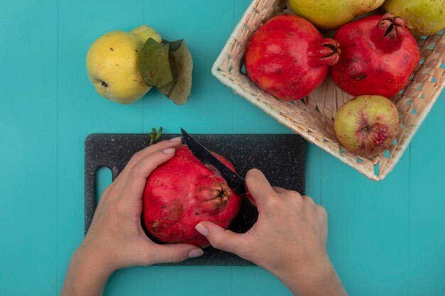 Top view of female hands cutting fresh pomegranate on a black kitchen board with knife on a blue background