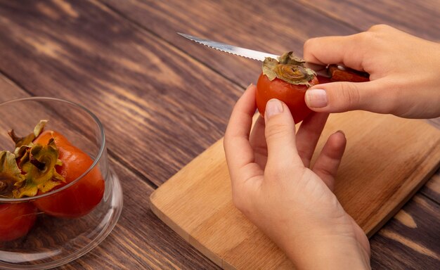 Top view of female hands cutting a fresh and soft persimmon on a wooden kitchen board with knife on a wooden surface