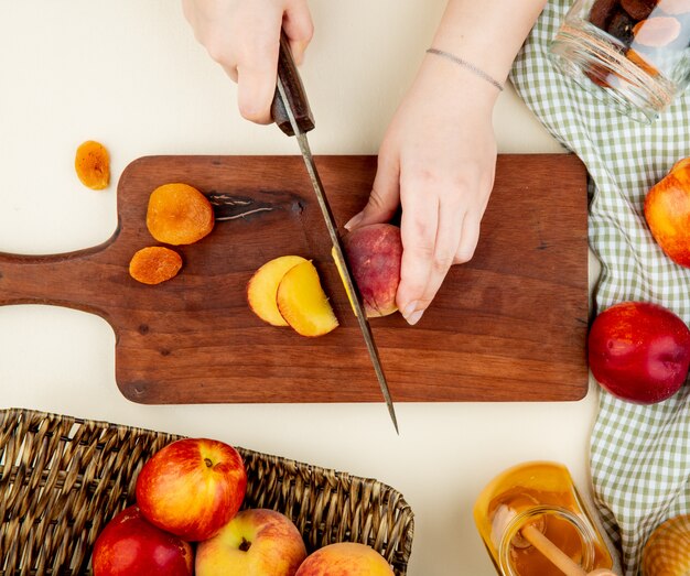 top view of female hands cutting fresh sweet peaches on a wooden cutting board on white