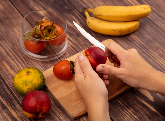 Top view of female hands cutting a juicy peach on a wooden kitchen board with knife with tangerine and bananas isolated on a wooden wall