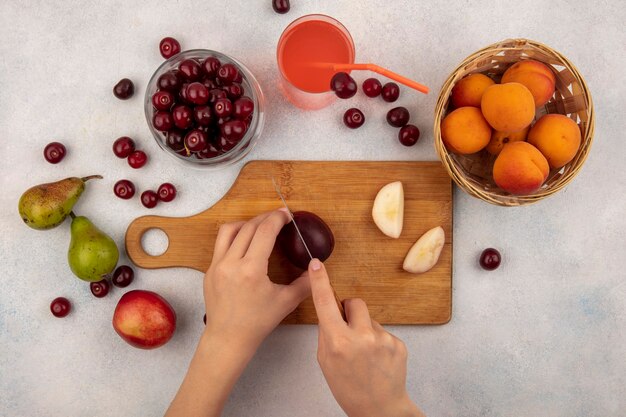Top view of female hands cutting peach with knife on cutting board and cherry juice with jar of cherry and basket of apricot with pears on white background