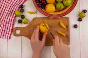 Free photo top view of female hands cutting peach on a wooden kitchen boar with knife with fruits like sloesgreen cherry plums isolated on a white wooden background