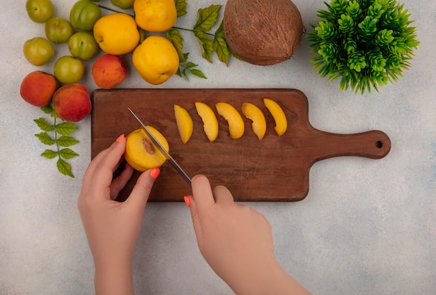 Foto gratuita vista dall'alto delle mani femminili che tagliano la pesca su una tavola di cucina in legno con prugne ciliegia verde su sfondo bianco