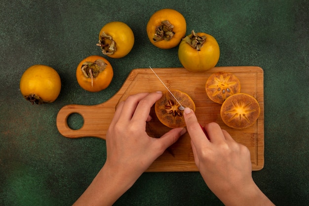 top view of female hands cutting persimmon fruits with knife on a wooden kitchen board on a green background