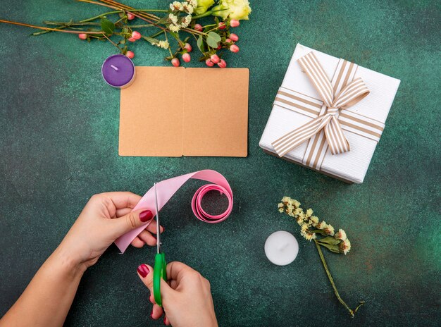 Top view of female hands cutting pink ribbon with yellow rose on gre with copy space