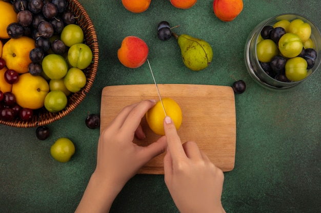 Top view of female hands cutting yellow peach with knife on a wooden kitchen board on a green background