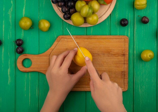 緑の木製の背景に木製キッチンボード上のナイフで黄色の桃を切る女性の手の上から見る
