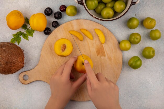 Top view of female hands cutting yellow peach on a wooden kitchen board with knife with coconut with peaches isolated on a white background