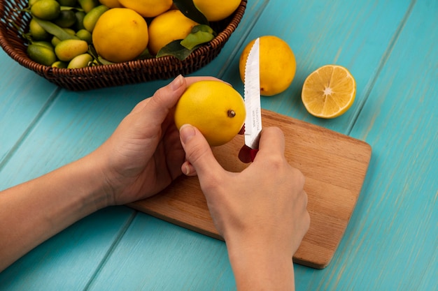 Top view of female hands cutting yellow skinned lemon on a wooden kitchen board with knife with fruits such as kinkans and lemons on a bucket on a blue wooden wall