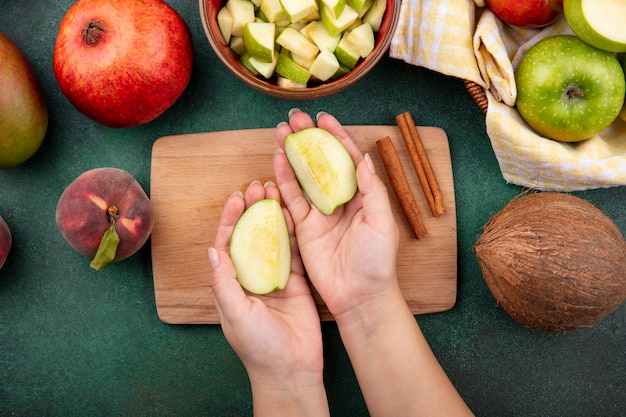 Вид сверху на женские руки, держащие яблоки над деревянной кухонной доской с палочками корицы и кокосом на зеленом