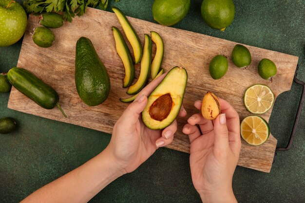 Top view of female hands holding an avocado in one hand and its pit in the other hand on a wooden kitchen board with limes feijoas and green apples isolated on a green surface