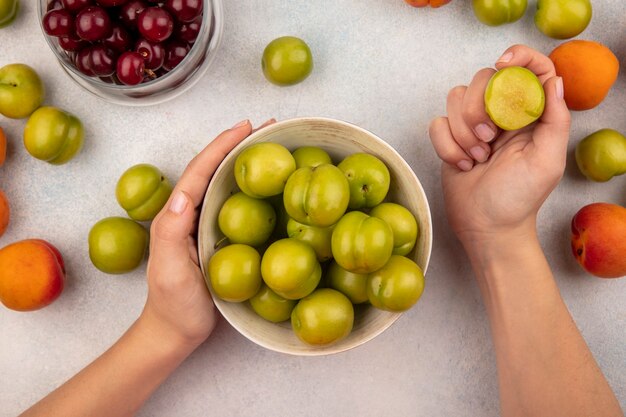 Top view of female hands holding basket of green plums and bitten plum with jar of cherries and apricots on white background