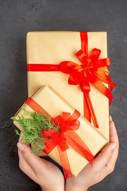 Top view female hands holding big and small xmas gifts in brown paper tied with red ribbon on dark background