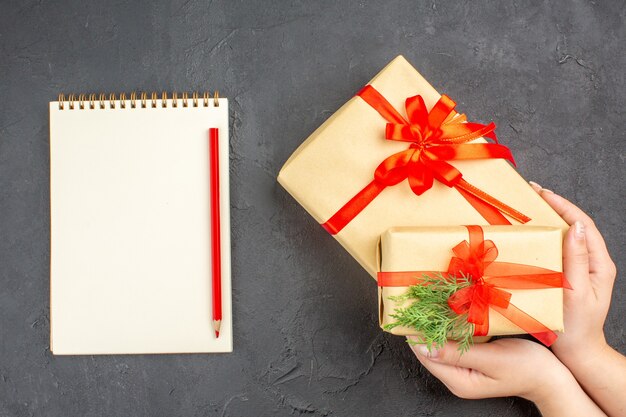 暗い表面に赤いリボンのノートブック鉛筆で結ばれた茶色の紙で大小のクリスマスプレゼントを保持している上面図の女性の手