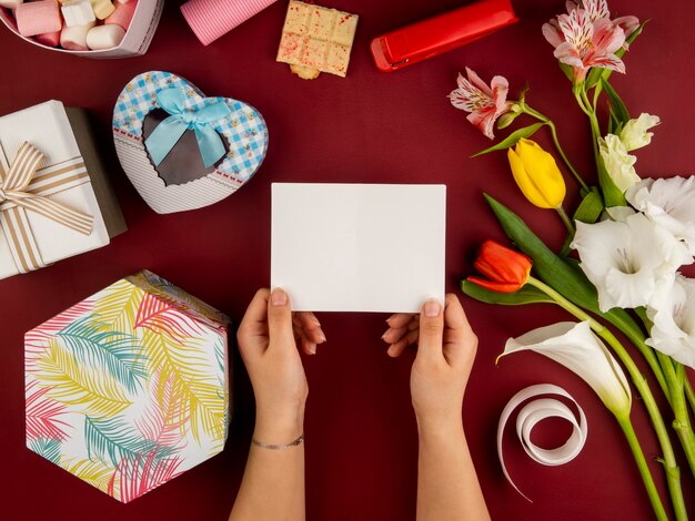 Top view of female hands holding blank paper greeting card over red table with red and yelow color tulips with alstroemeria and heart shaped gift box and white chocolate