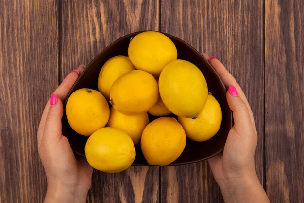 Top view of female hands holding a bowl of fresh lemons on a wooden wall