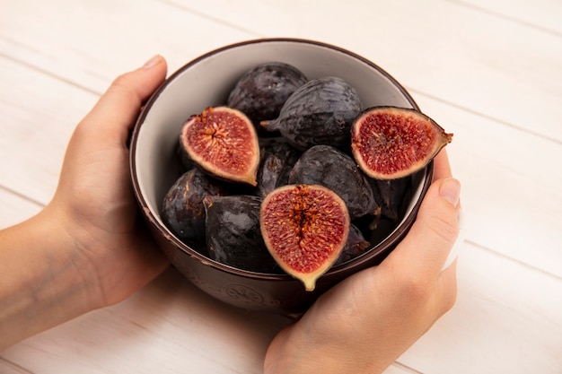 Top view of female hands holding a bowl of fresh sweet black mission figs on a white wooden wall