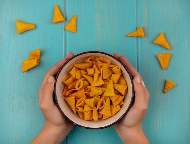 Top view of female hands holding a bowl of tasty cone shape corn snacks on a blue wooden table