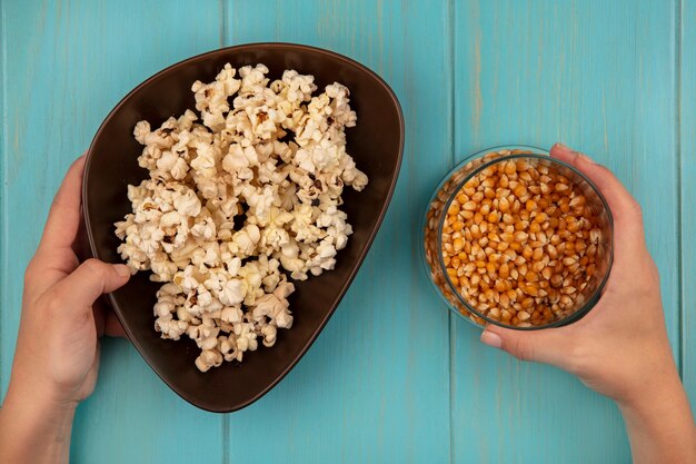 Top view of female hands holding a bowl of tasty popcorns in one hand and in the other hand a bowl of popcorn kernels on a blue wooden table