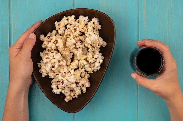 Vista dall'alto delle mani femminili che tengono una ciotola di gustosi popcorn in una mano e nell'altra un bicchiere di cola su un tavolo di legno blu