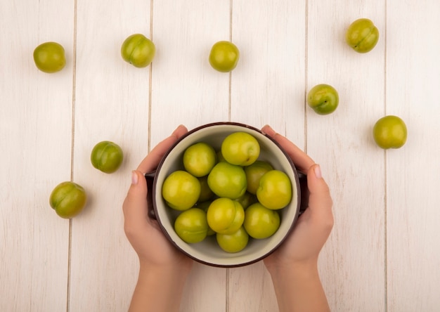 Top view of female hands holding a bowl with green cherry plum on a white wooden background